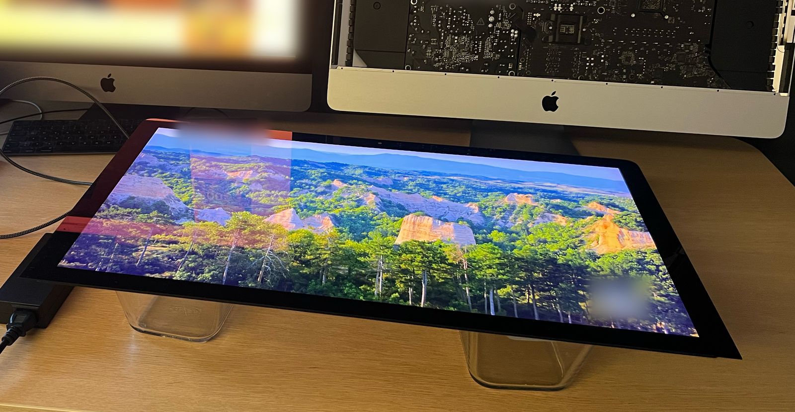 5K iMac monitor conversion in progress: the bare 27-inch 5K panel of an iMac lying on two clear stands on a desk, lit up by the driver board and showing a landscape of forest and rock formations. The board's black power adapter sits at the left edge; behind the panel stands the opened iMac with its internal parts exposed.