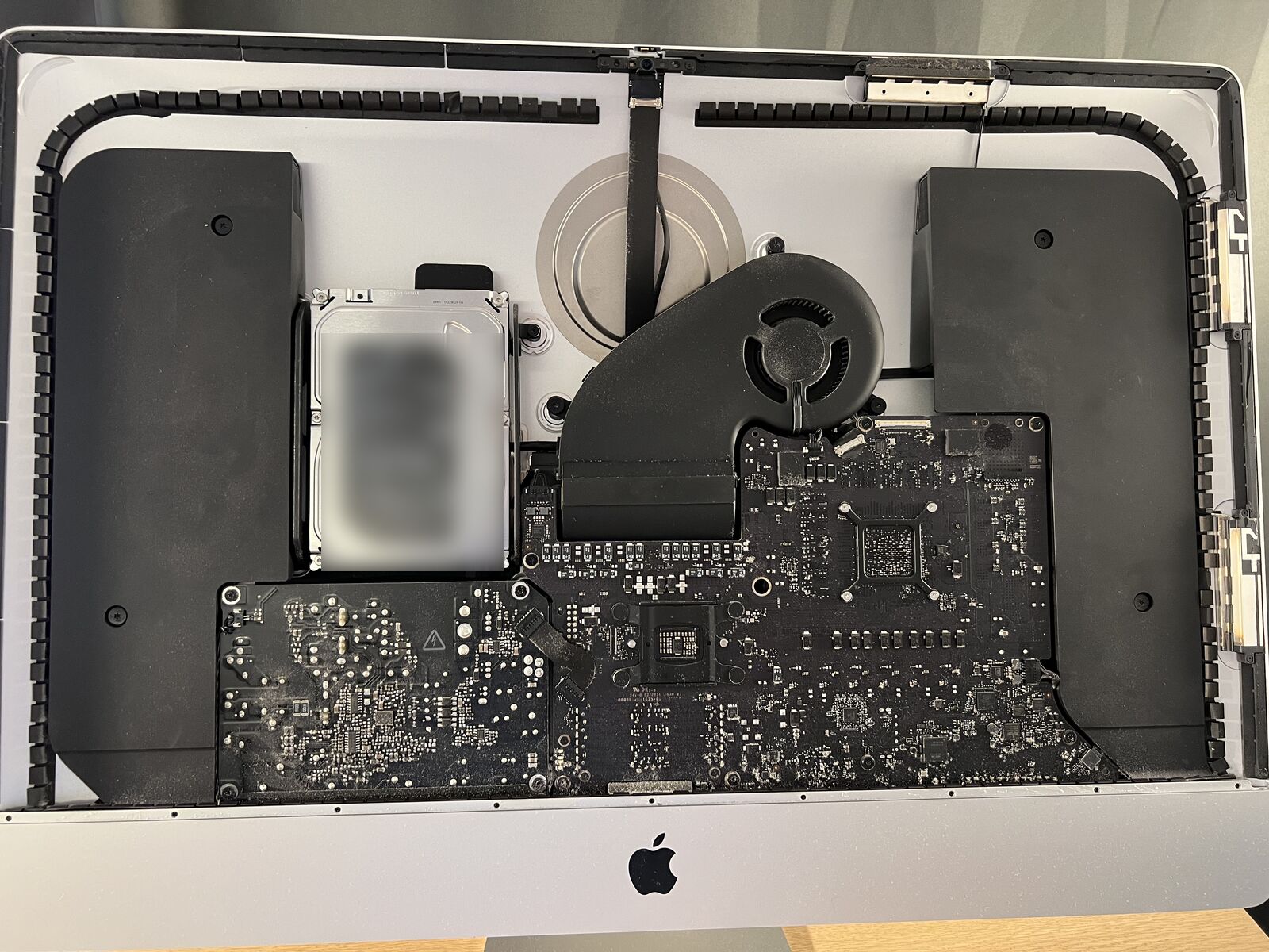 A 27-inch 5K iMac lying face up with the display panel removed: the power supply with a warning triangle at the bottom left, the logic board at the bottom right, the fan, the hard drive and the two speakers above.