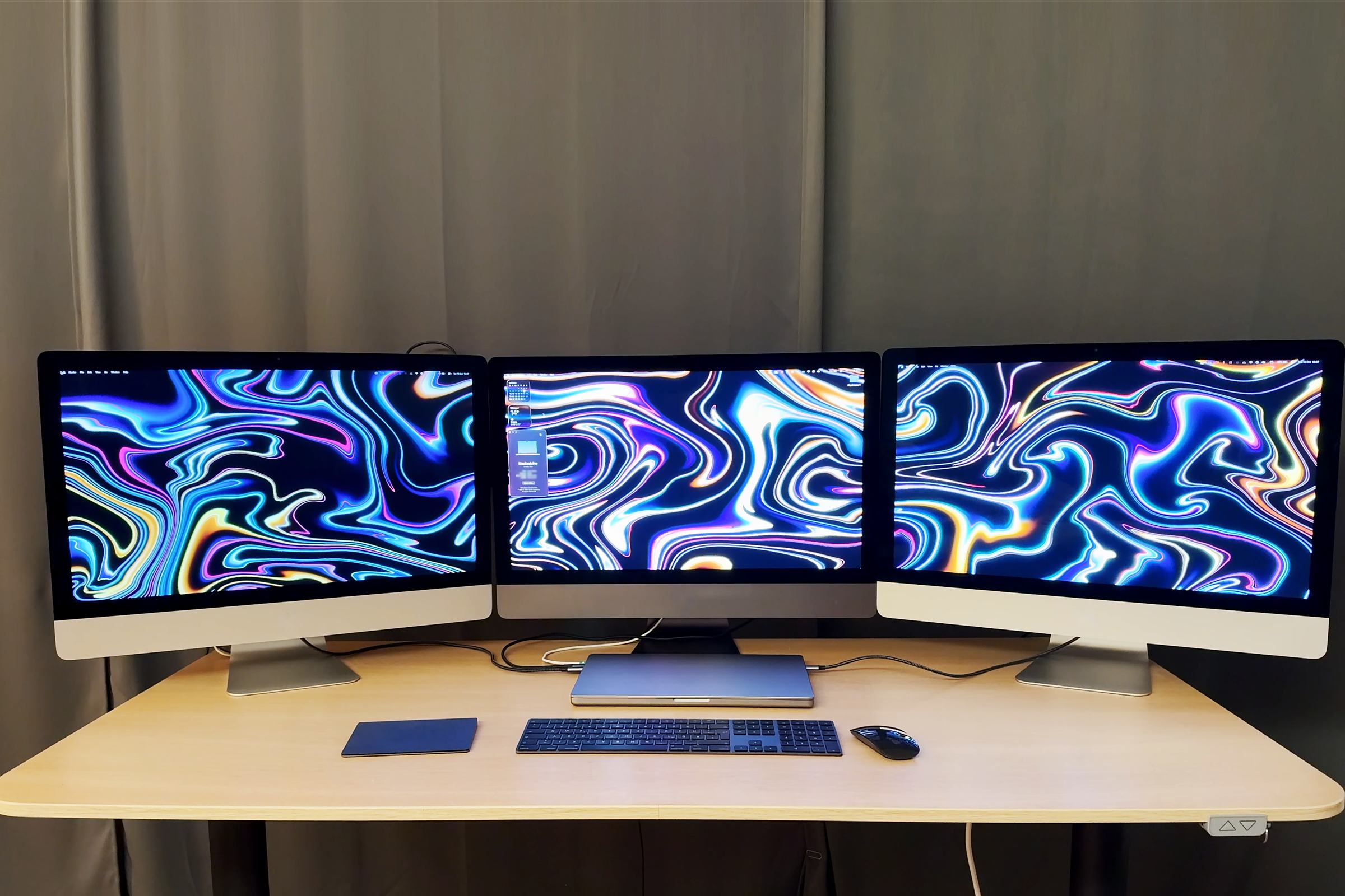 Três iMacs de 27 polegadas lado a lado com um papel de parede em espiral que atravessa as três telas, funcionando como telas do MacBook Pro fechado na mesa à frente deles, conectado por cabos.