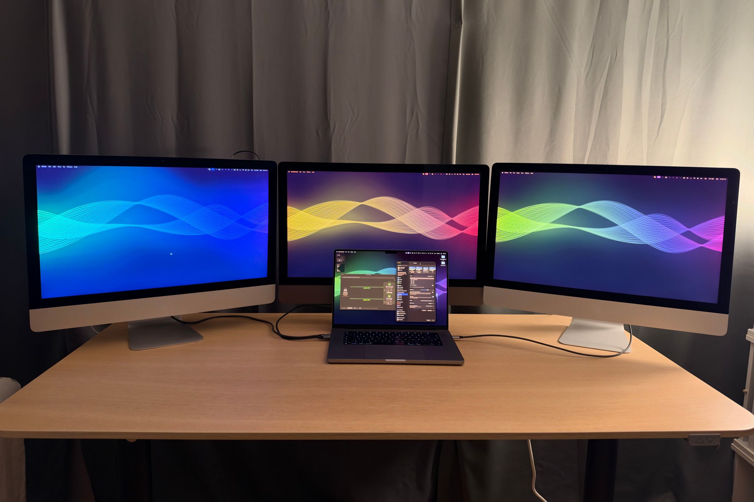 Tres iMac de 27 pulgadas con fondos de ondas de colores como pantallas extendidas del MacBook Pro que está delante en el escritorio.