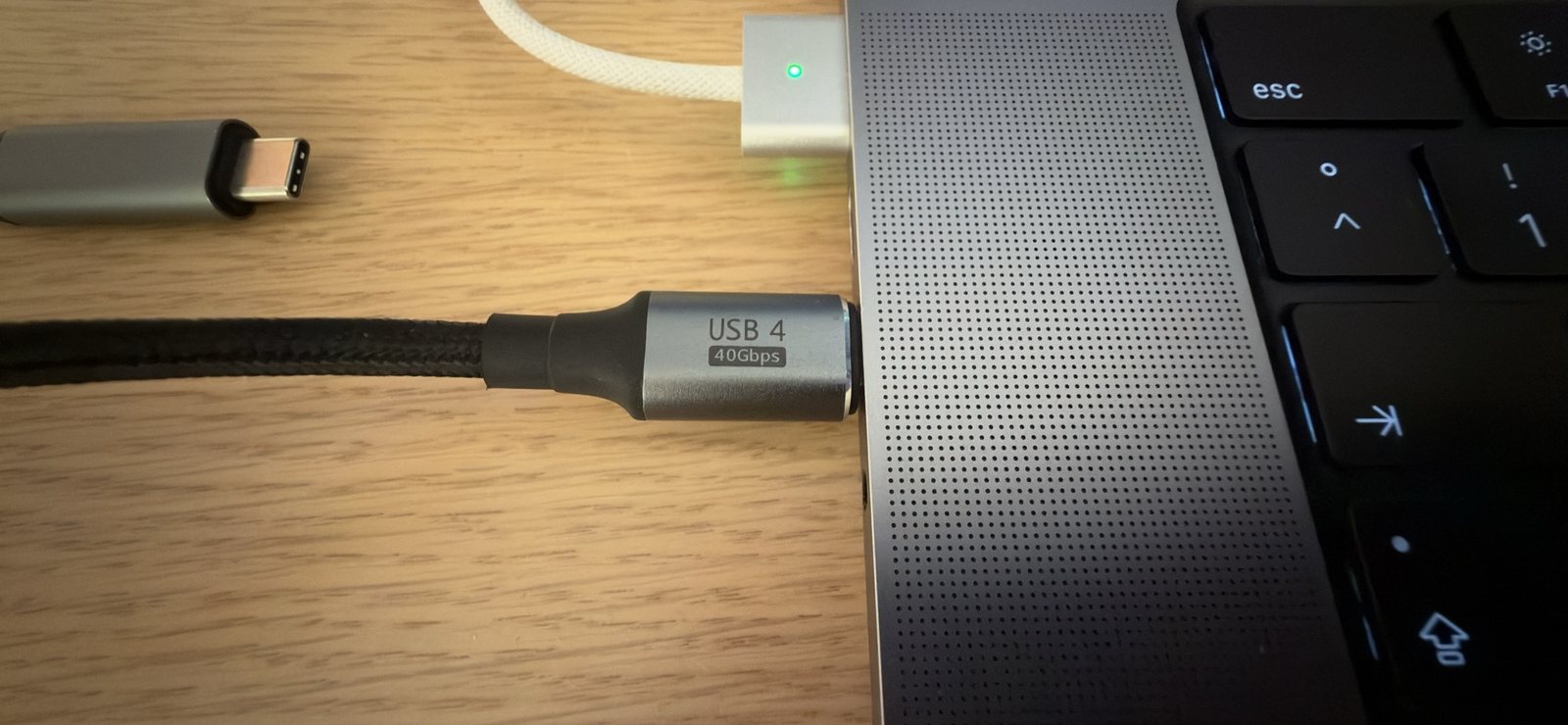 Close de um cabo preto trançado conectado ao lado esquerdo de um MacBook Pro; o conector prateado traz a inscrição USB 4, 40Gbps. Acima dele, um carregador MagSafe está conectado.